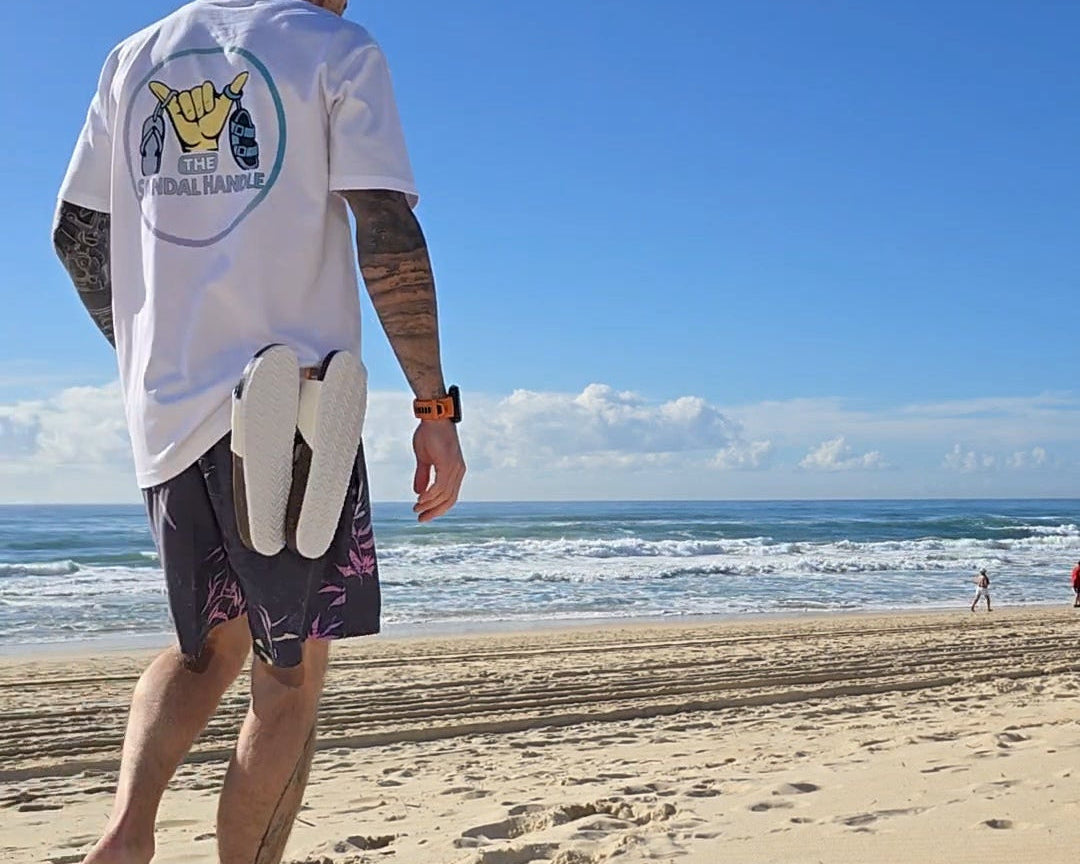 A man standing on a sandy beach facing the ocean, wearing a white graphic t-shirt with a yellow and blue logo on the back, dark patterned swim trunks, and sunglasses.