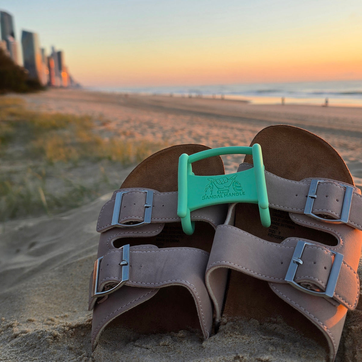 A pair of brown double-strap sandals with silver buckles and a mint green Sandal Handle, resting on a sandy beach with a sunset sky and ocean in the background.