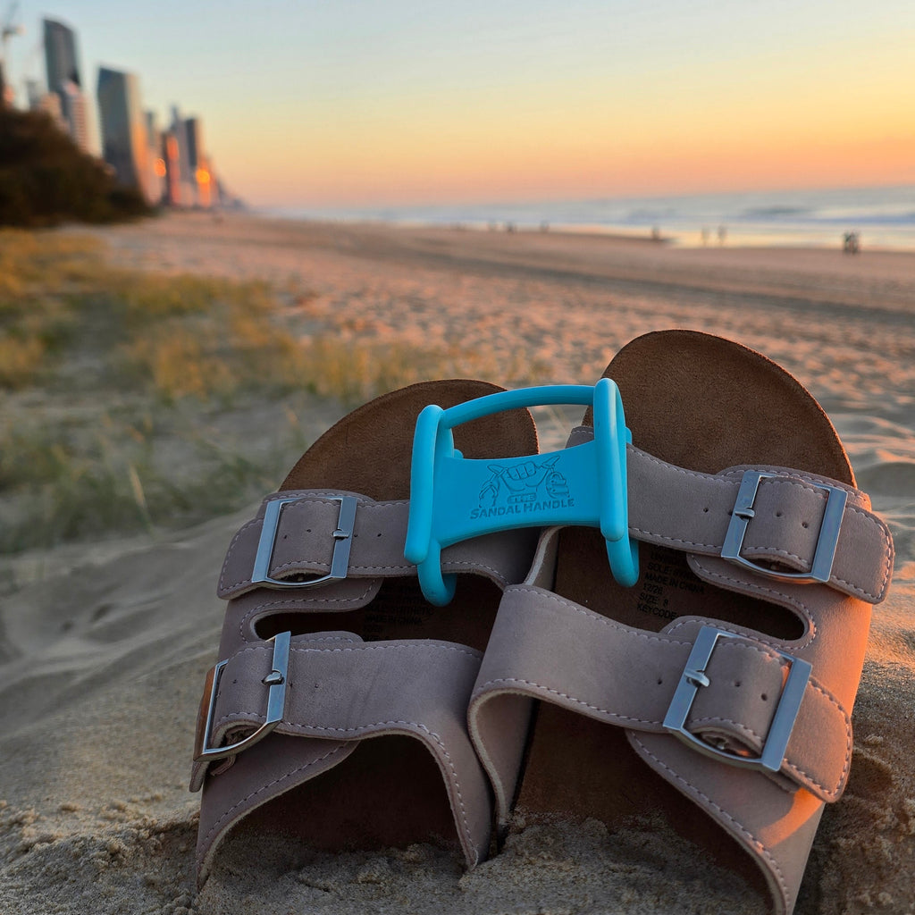A pair of brown leather sandals with adjustable straps and a light blue Sandal Handle, placed on sand at a beach during sunset with a city skyline in the background.