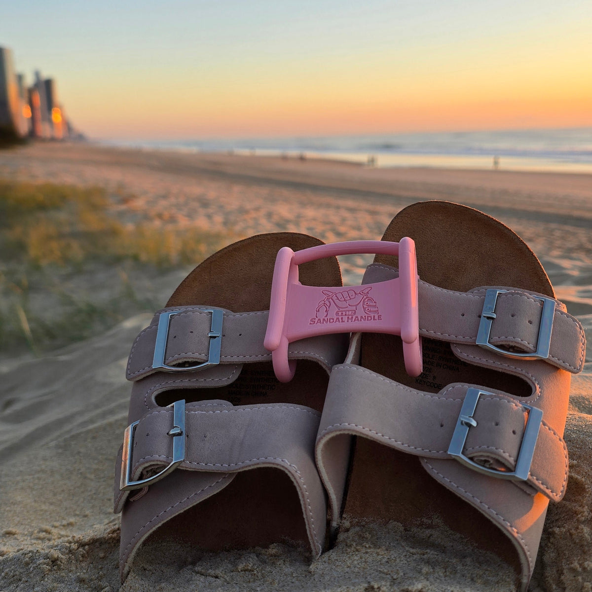 A pair of brown leather double-strap sandals with silver buckles and a peach pink Sandal Handle, placed on a sandy beach with a sunset ocean view in the background.