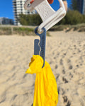 A hand holds a white plastic bag clip with a gray metal clip holding a dog poo bag set against a blurred beach background with sand, dunes, and buildings.