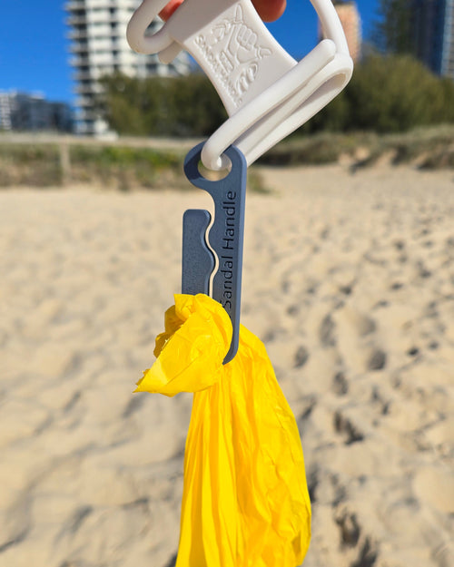 A hand holds a white plastic bag clip with a gray metal clip holding a dog poo bag set against a blurred beach background with sand, dunes, and buildings.