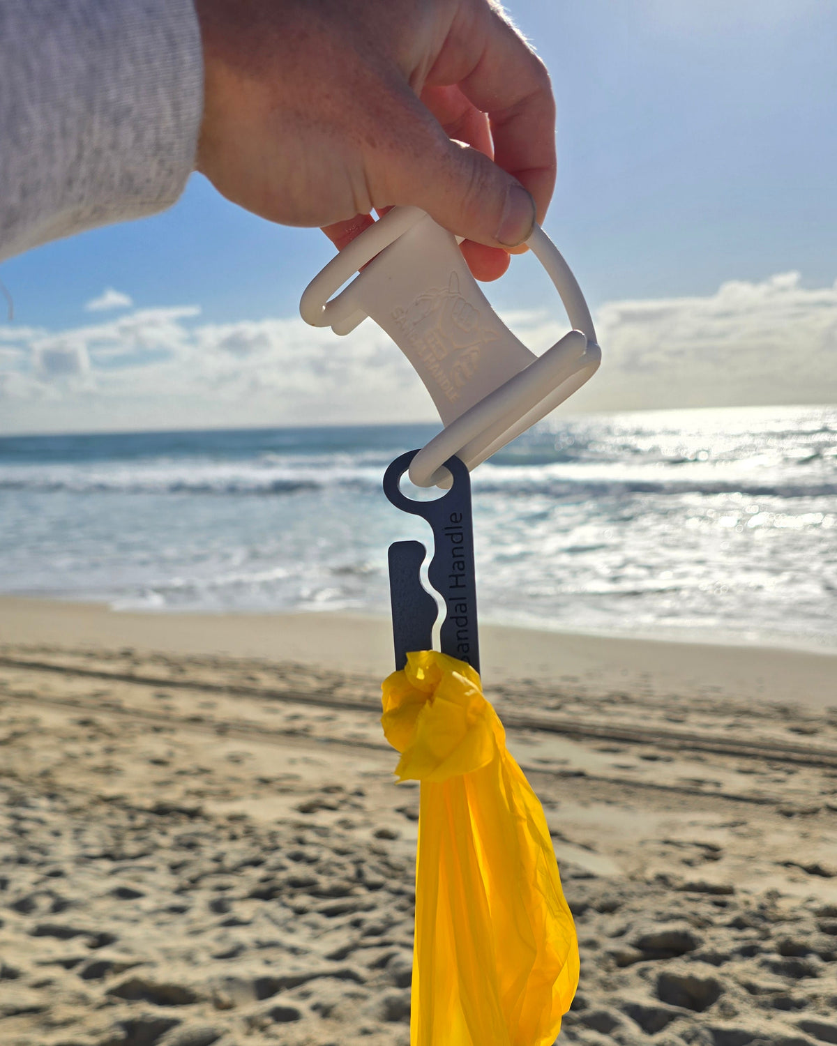 A hand holding a white plastic bag opener tool with a yellow reusable bag attached, photographed over a sandy beach with the ocean in the background.