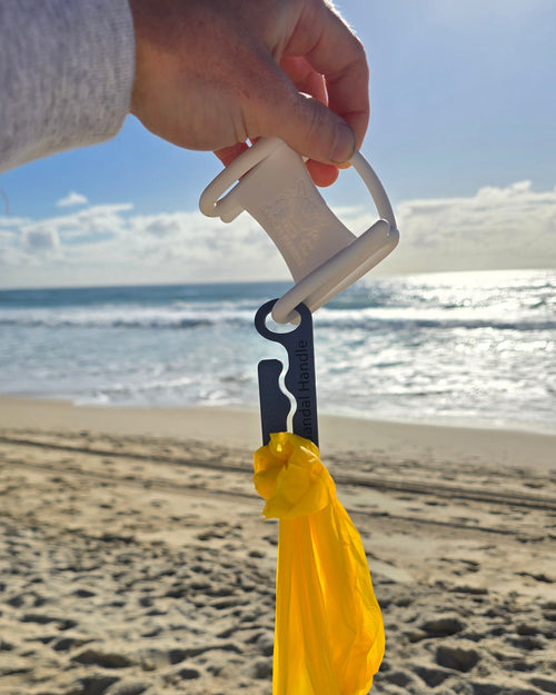 A hand holding a white plastic bag opener tool with a yellow reusable bag attached, photographed over a sandy beach with the ocean in the background.